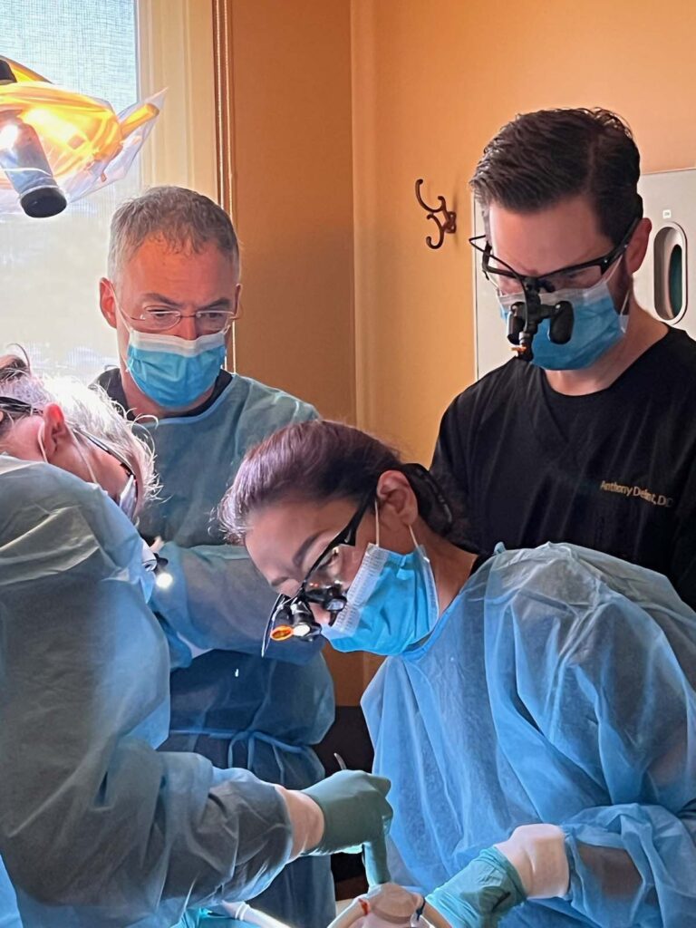 Four medical professionals in surgical gowns, gloves, and masks, with a surgical light overhead, engaged in a medical procedure.