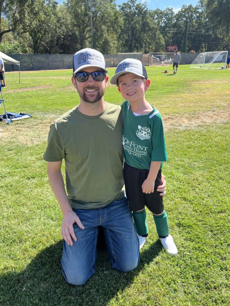Adult and child in soccer uniforms posing on a grassy field, with soccer goals and other people visible in the background.