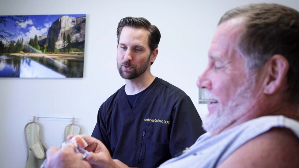 The image shows a dental office setting with a person wearing a black dental uniform that has 'Anthony DeFont, DDS' embroidered on it. The person is holding a dental model, likely explaining something to a patient.