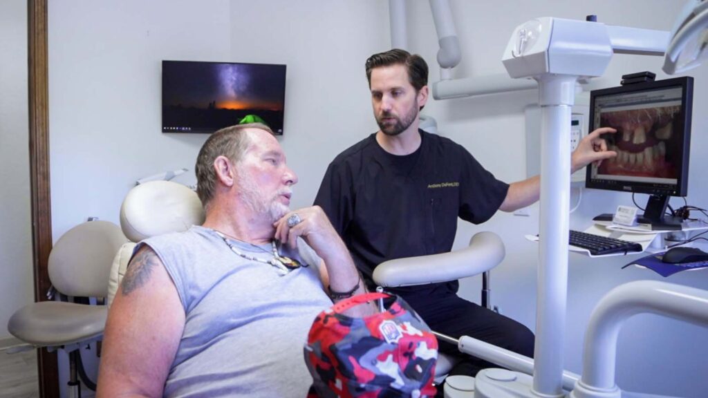 A dentist wearing a black uniform with 'Anthony DeFont, DDS' embroidered on it, sitting next to a patient in a dental chair, looking at a computer monitor in a dental office setting.